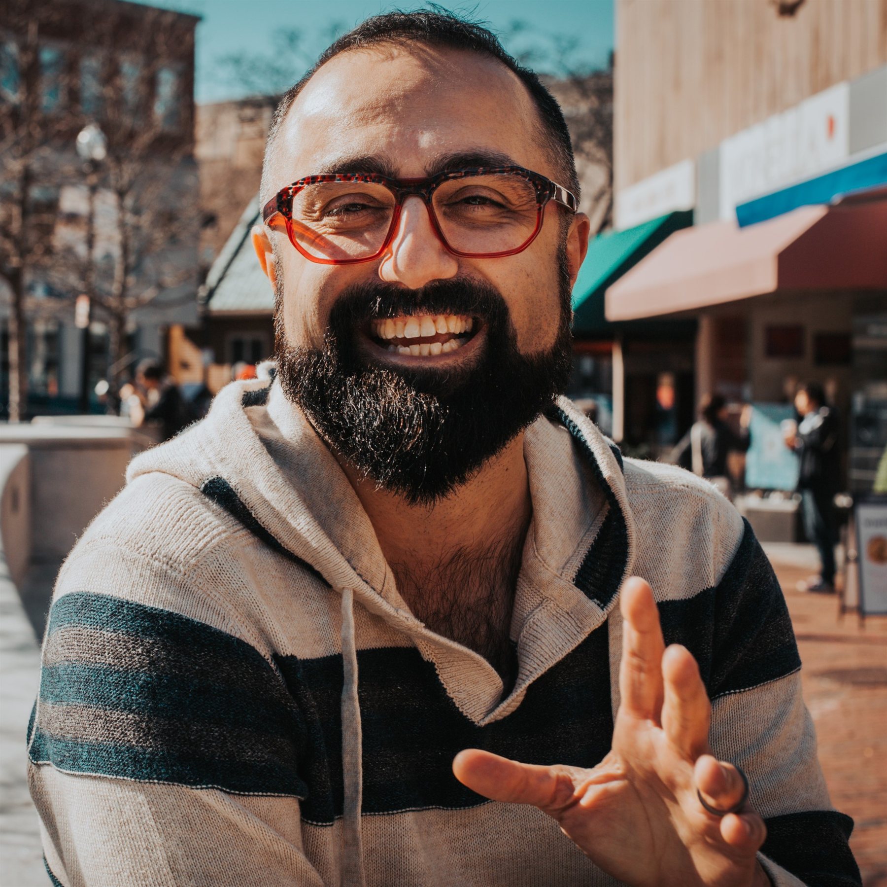 Andy in a striped hooded sweater smiles and gestures toward the camera with one hand; storefronts line the street behind him.