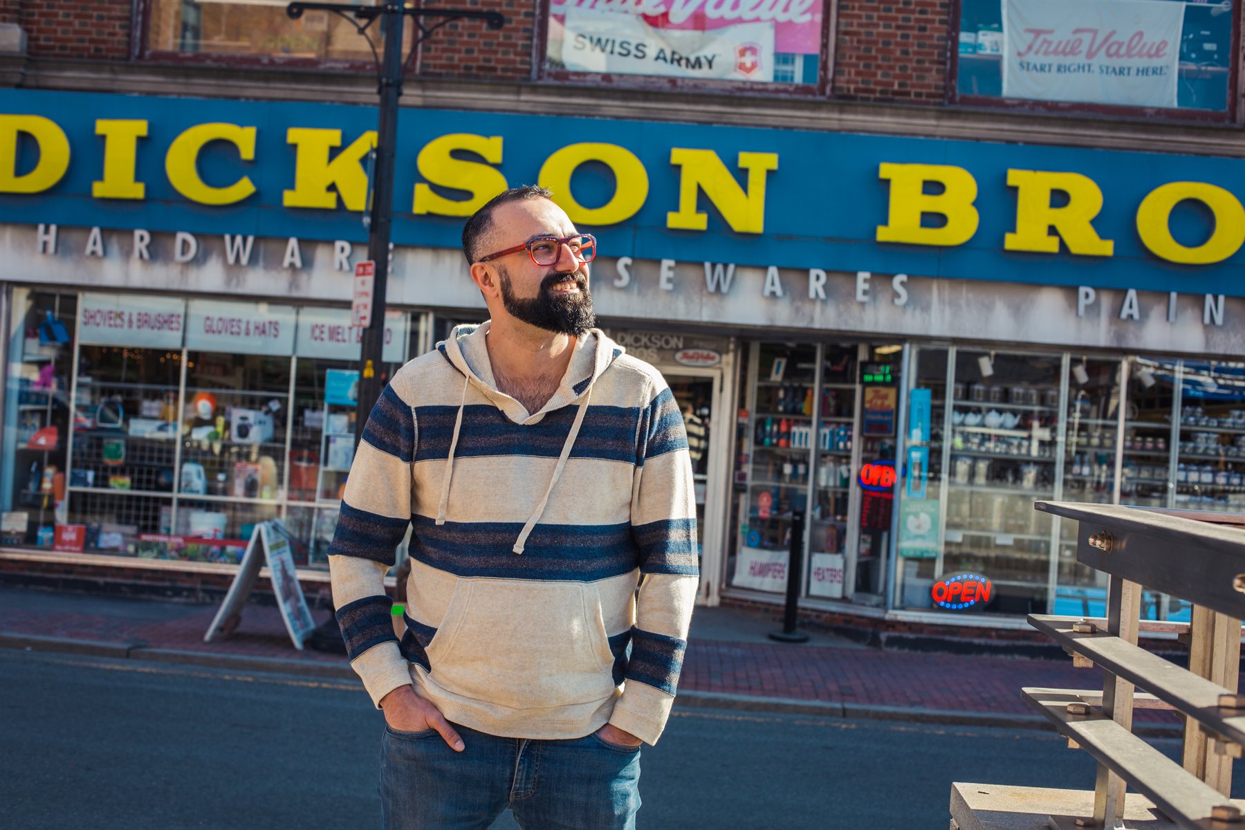 Andy stands on a city sidewalk in a striped hoodie, with the Dickson Bros. storefront sign behind him.