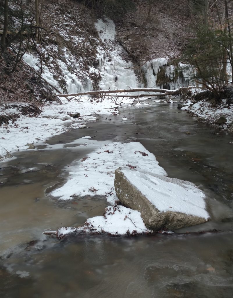 A hiker crosses a partly frozen stream by stepping across icy rocks.